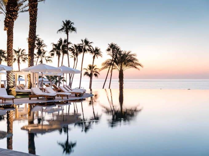 Luxury Resort Pool With White Sunbeds And Umbrellas, Overlooking Palm Trees And The Ocean At Sunset In Puerto Los Cabos, Baja California Sur, Mexico, For Real Estate And Vacation Rental Opportunities.