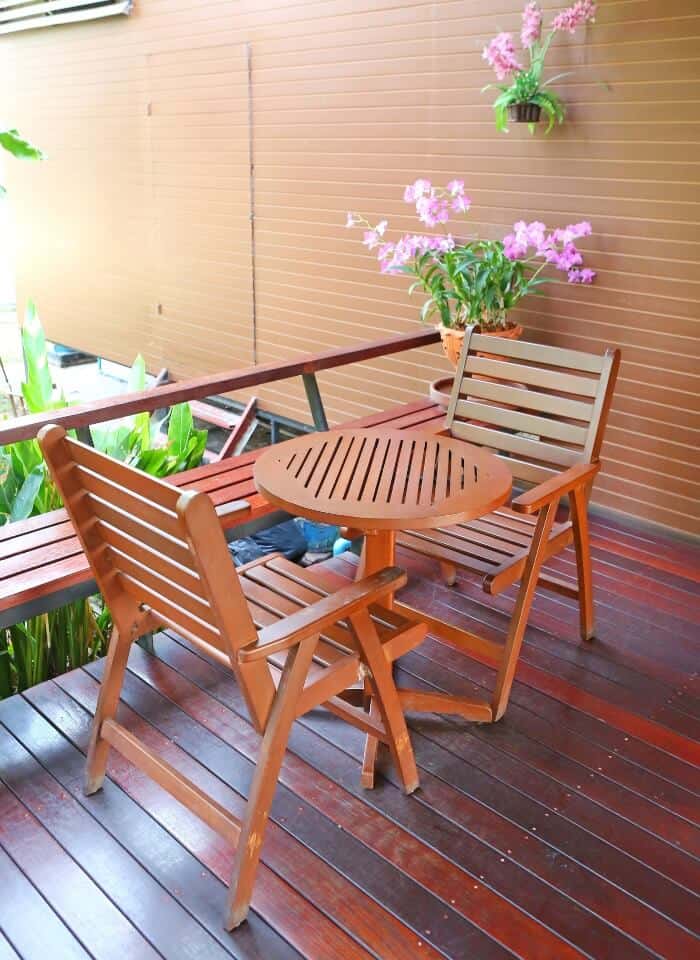 A small outdoor patio features a round wooden table with two matching wooden chairs. Pink flowers hang on the wooden wall behind, and green plants surround the cozy seating area.