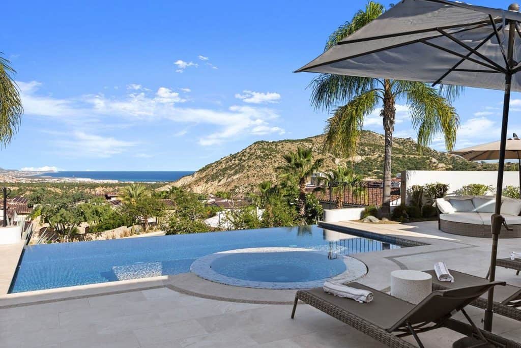 Stunning Querencia Real Estate Outdoor Pool Area With Mountain And Ocean Views, Palm Trees, Lounge Chairs, And Umbrellas In A Luxurious Setting.