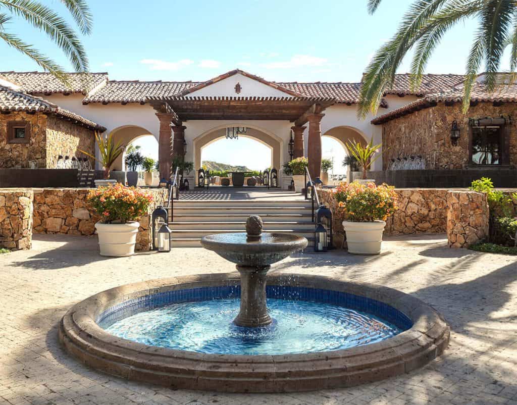 Luxury Mediterranean-Style Estate Entrance With Stone Accents And A Central Fountain, Surrounded By Lush Potted Plants And Palm Trees, Embodying Querencia'S Upscale Real Estate Charm.