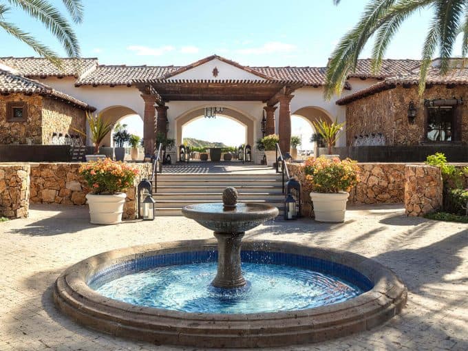 Luxury Mediterranean-Style Estate Entrance With Stone Accents And A Central Fountain, Surrounded By Lush Potted Plants And Palm Trees, Embodying Querencia'S Upscale Real Estate Charm.