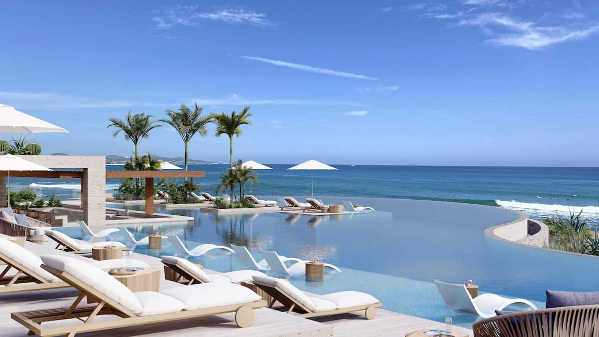 Aerial View Of A Luxurious Infinity Pool With Lounge Chairs, Palm Trees, And Ocean Views At Querencia Real Estate. Perfect For Relaxation And Enjoying The Stunning Coastal Scenery.