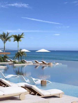 Aerial View Of A Luxurious Infinity Pool With Lounge Chairs, Palm Trees, And Ocean Views At Querencia Real Estate. Perfect For Relaxation And Enjoying The Stunning Coastal Scenery.