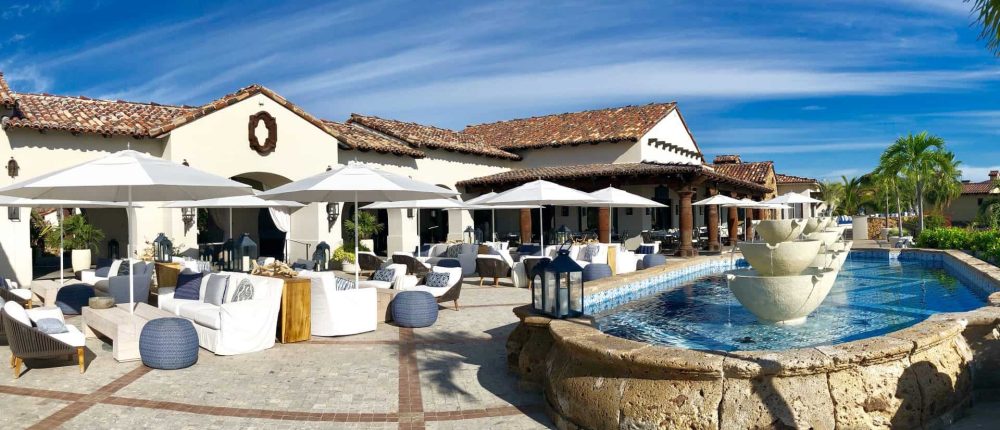 Elegant Querencia Luxury Estate With A Spacious Outdoor Lounge Area, Poolside Fountains, And Lush Tropical Landscaping Under A Bright Blue Sky.