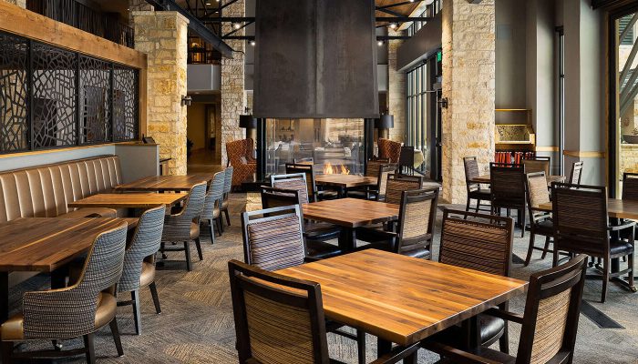 Modern Dining Area At Querencia Featuring Warm Wood Furniture, A Cozy Fireplace, And Large Glass Windows With Natural Light, Perfect For A Luxurious Living Experience.