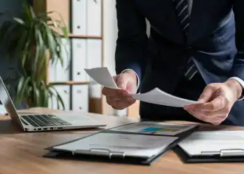 Profissional de neg&oacute;cios em traje azul analisa documentos financeiros em uma mesa de madeira com um laptop aberto.