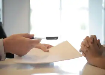 Imagem de uma pessoa segurando uma caneta e um contrato. Em sua frente, uma outra pessoa com as m&atilde;os entrela&ccedil;adas.