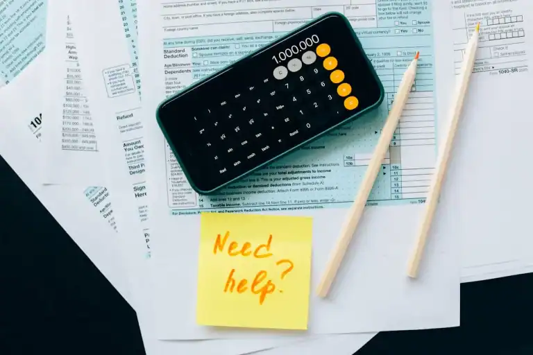A imagem mostra um celular aberto na calculadora, papéis de documentos, talvez recibos ou formulários, sugerindo atividades como cálculo de impostos, análise fiscal ou declaração
