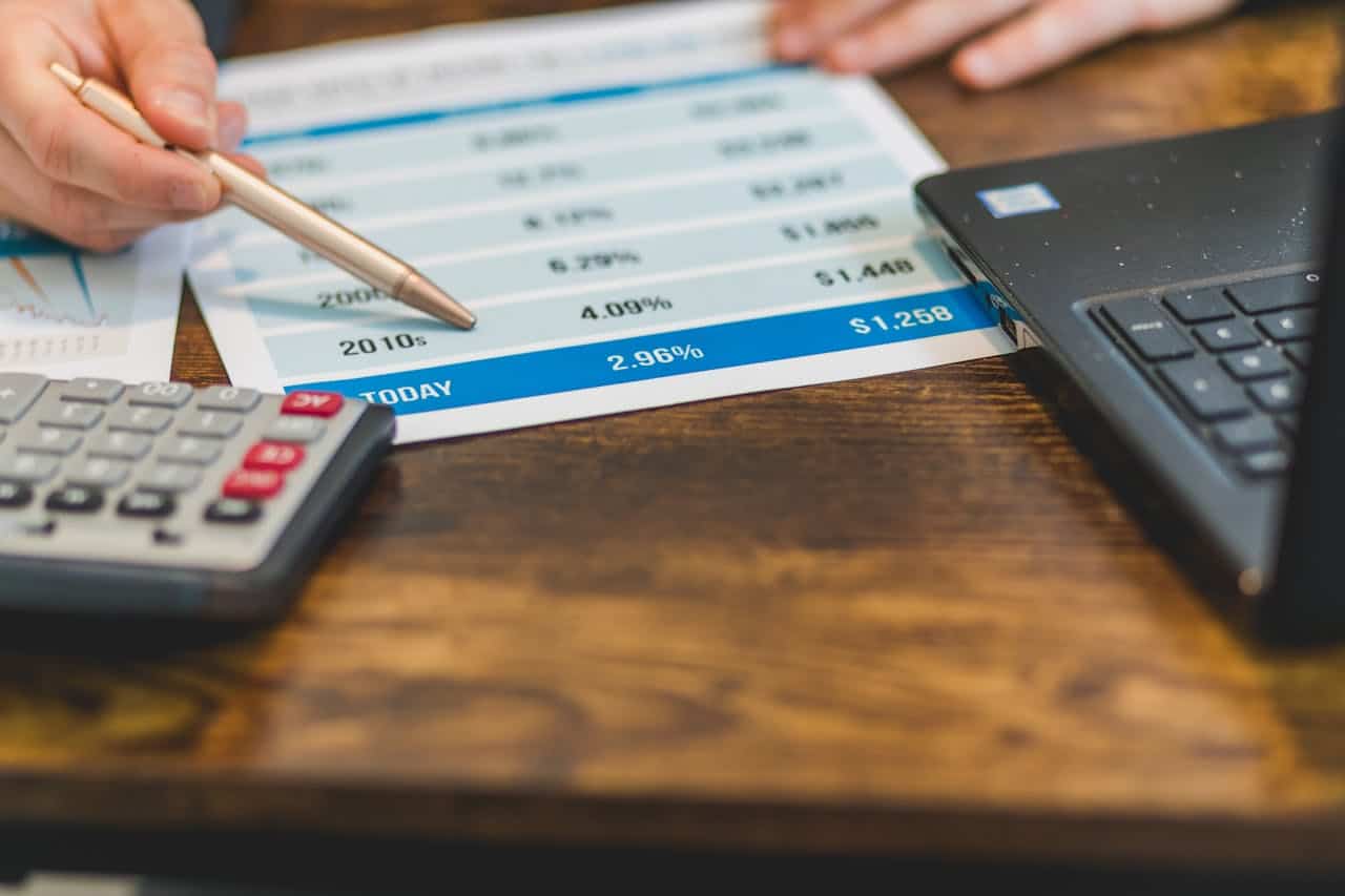 A imagem mostra uma mesa de trabalho organizada, com um papel contendo uma tabela financeira no centro, uma calculadora ao lado e um notebook aberto próximo, sugerindo análise de dados ou planejamento financeiro.