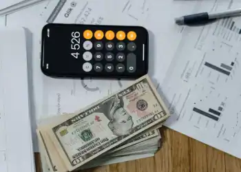 Imagem mostra um celular com a calculadora aberta, ao lado de algumas notas de dinheiro sobre pap&eacute;is com gr&aacute;ficos.