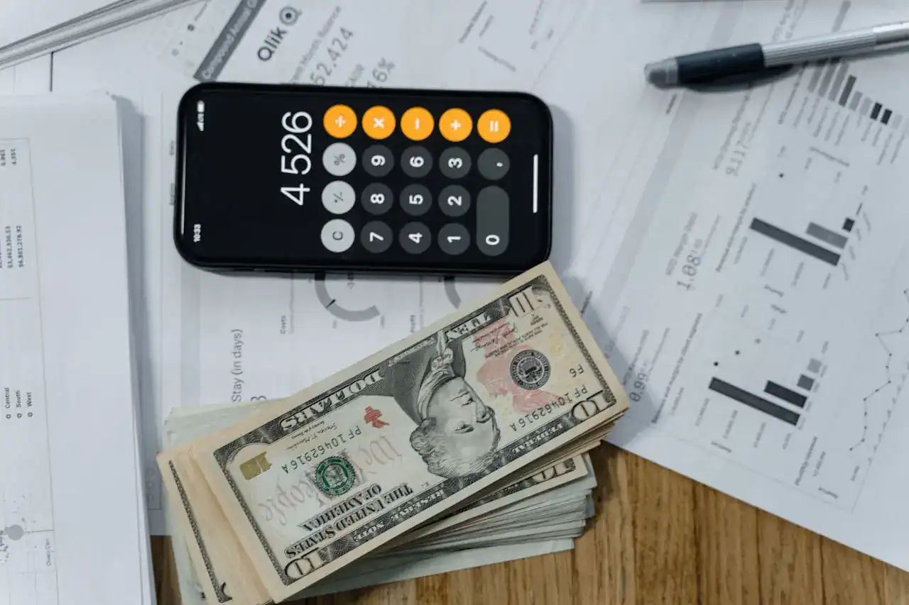 Imagem mostra um celular com a calculadora aberta, ao lado de algumas notas de dinheiro sobre papéis com gráficos.