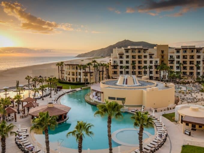 Luxurious Quivira Los Cabos Resort With An Oceanfront Pool And Stunning Sunset View, Surrounded By Palm Trees And Modern Architecture, Offering The Perfect Beach Vacation Experience.