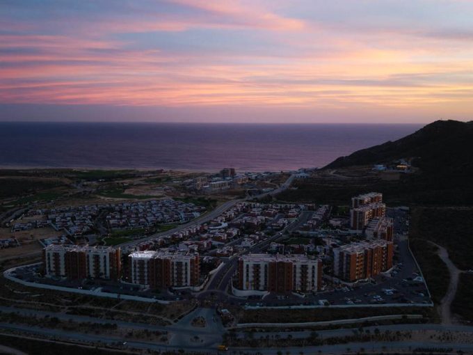 Stunning Aerial View Of Quivira Los Cabos Real Estate With Colorful Sunset Sky, Overlooking Luxurious Residential Buildings, Scenic Hills, And The Pacific Ocean, Highlighting Luxurious Coastal Living.