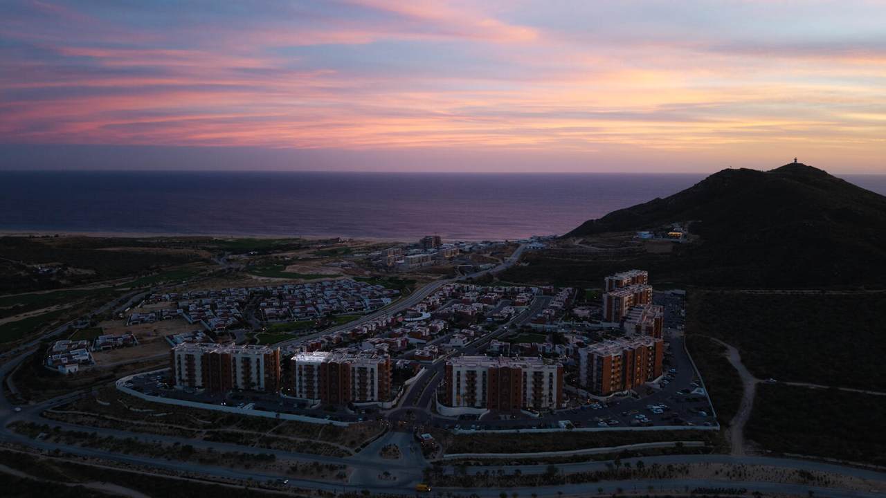 Stunning Aerial View Of Quivira Los Cabos Real Estate With Colorful Sunset Sky, Overlooking Luxurious Residential Buildings, Scenic Hills, And The Pacific Ocean, Highlighting Luxurious Coastal Living.