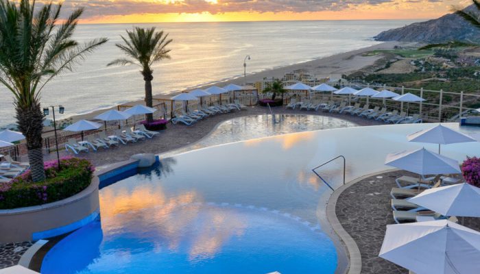 Wide View Of A Stunning Quivira Infinity Pool And Deck With White Umbrellas, Reflecting A Vibrant Sunset Over The Pacific Ocean.