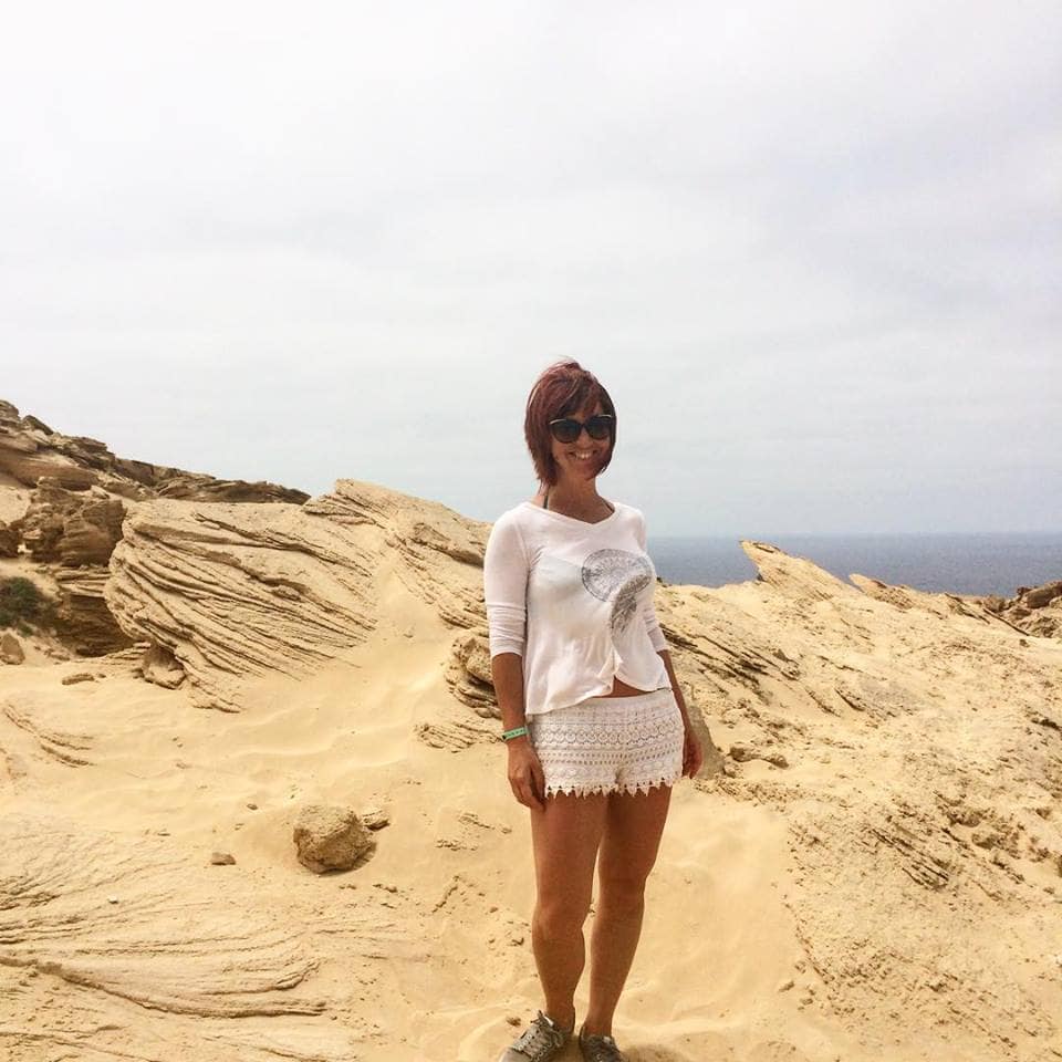 Beach landscape with a woman in a white dress and sunglasses standing on sandy rocks.