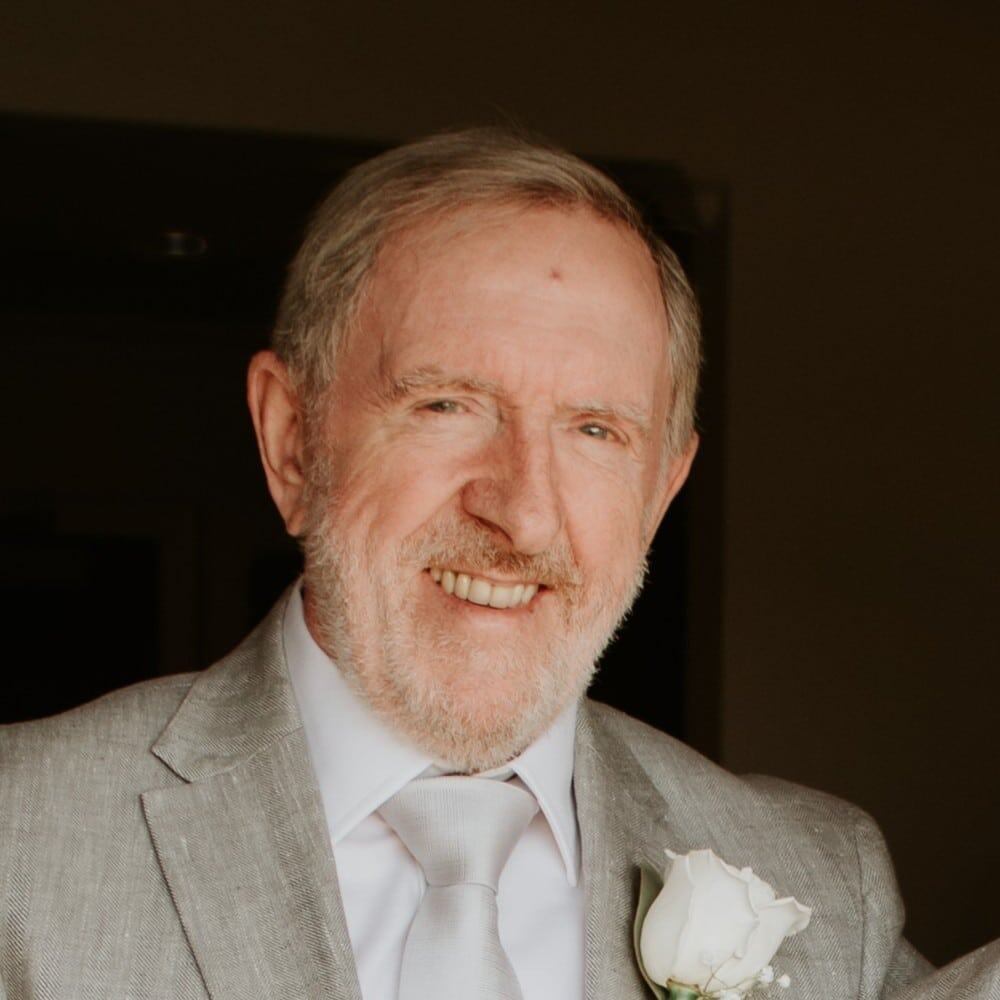 Senior man in a gray suit with a white tie, smiling warmly at a celebration or event.