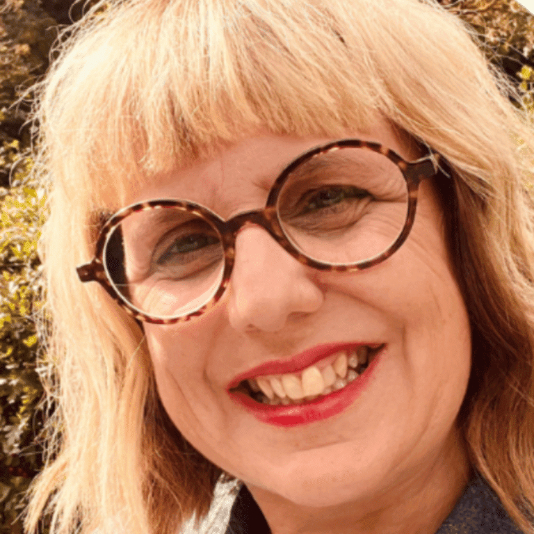 Woman with blonde hair and glasses smiling outdoors, natural sunlight.