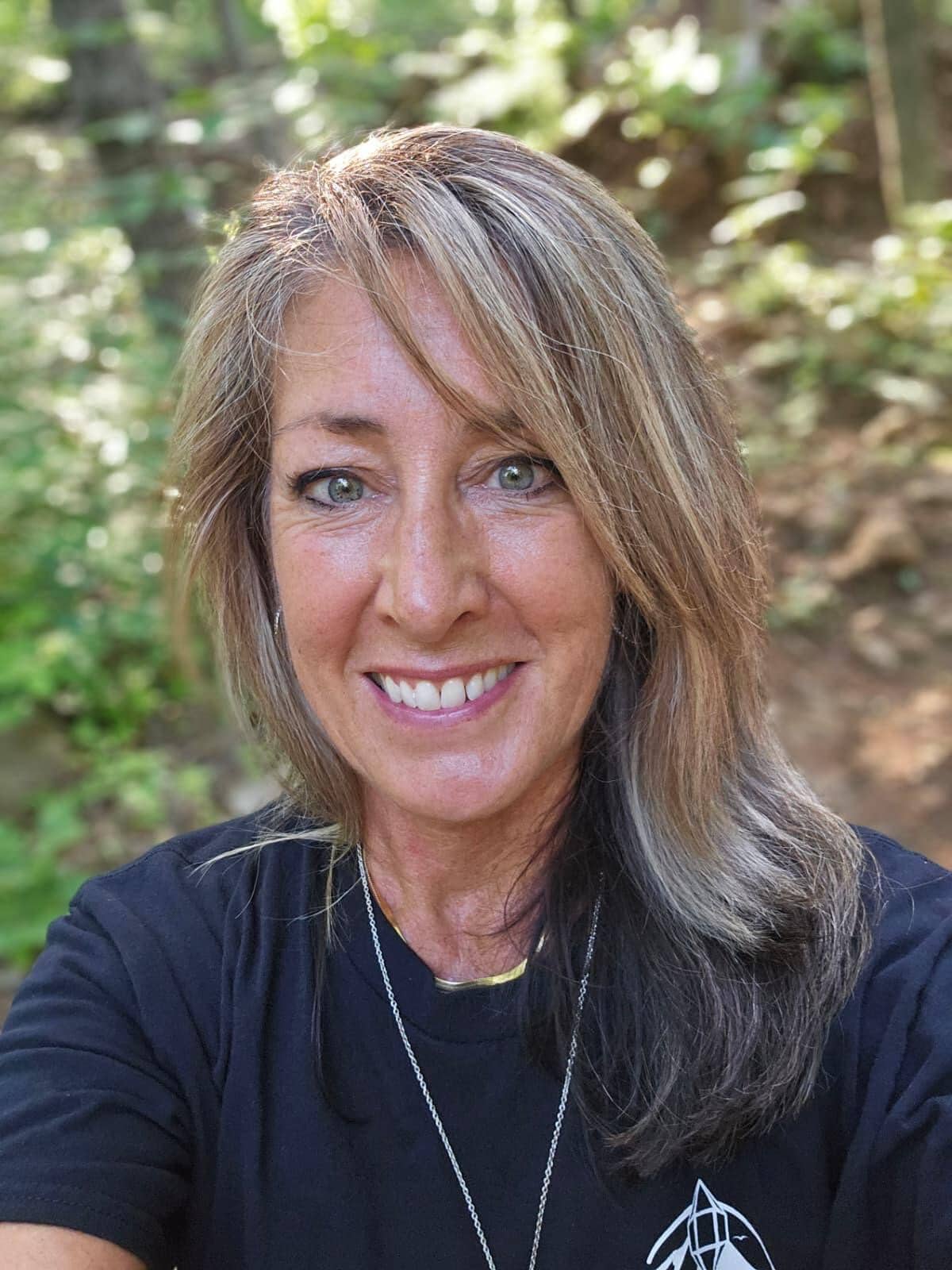 A woman smiling outdoors in a natural setting with greenery in the background.