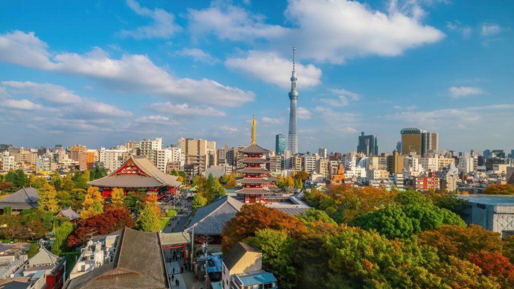 Tokyo skyline with a red pagoda and temple grounds in foreground, and the Skytree tower rising above a mix of city buildings under a blue sky.
