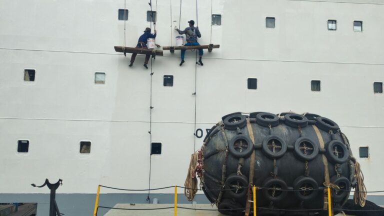 Two workers on a suspended platform clean or repair the white hull of a large ship, buckets in hand and harnessed for safety.