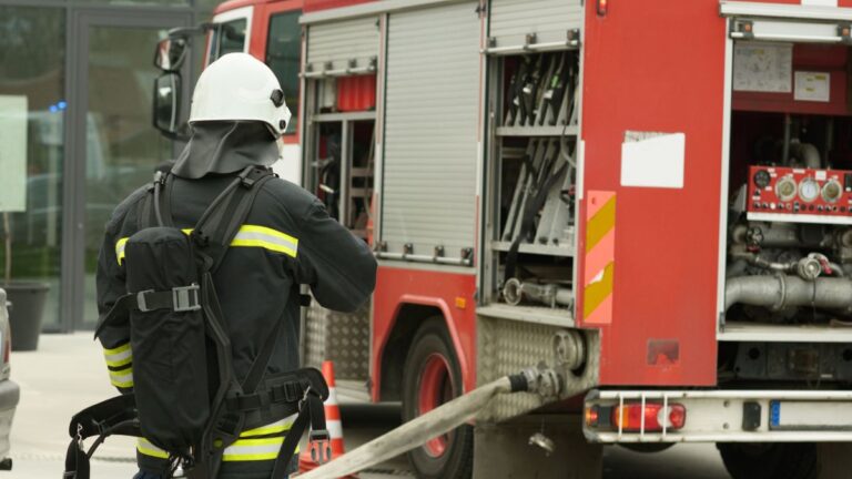 Firefighter in full turnout gear and helmet standing beside a red fire truck with a hose connected to the pump.