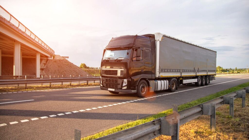 Black semi-truck driving on a highway with an overpass under construction to the left, sunset glow nearby