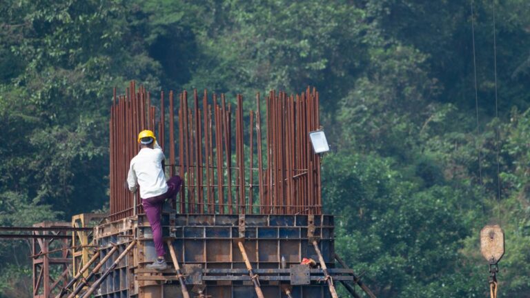 Construction worker in a yellow helmet climbs a rusted rebar framework at a construction site with trees in the background, wearing a white shirt and purple pants in mid-climb.