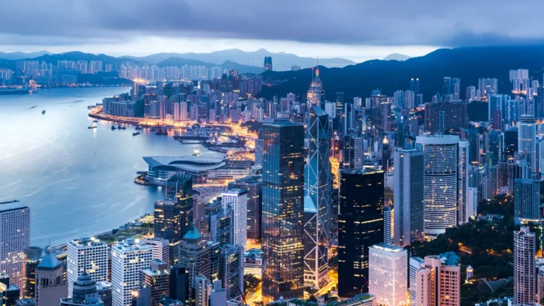 Evening city skyline of Hong Kong with tall skyscrapers along Victoria Harbour and mountains in the background.