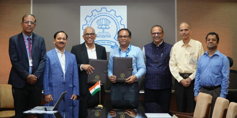 Seven professionals in a conference room pose for a formal agreement signing, two center figures holding official folders with an Indian flag on the table and a crest in the background.