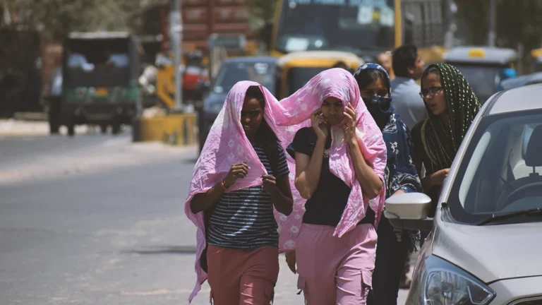 Three young women walk along a busy street with pink scarves draped over their heads; one looks down at a phone while the others adjust their scarves as cars pass by in the background.