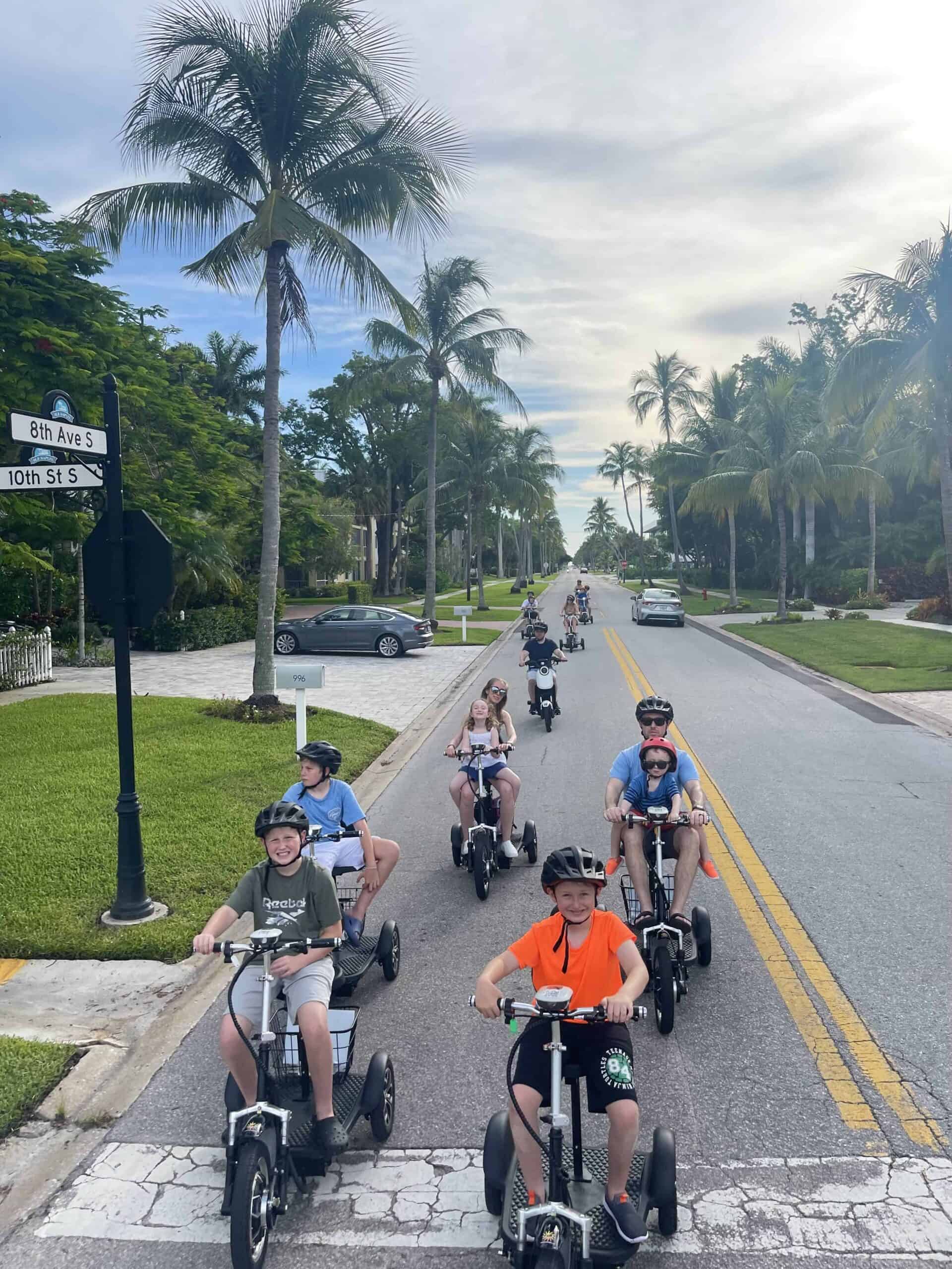 Children riding pedal bikes and tricycles on a sunny street in downtown Naples, FL, surrounded by palm trees and lush greenery. The group appears to be enjoying a fun bike tour or rental outing in a scenic area.
