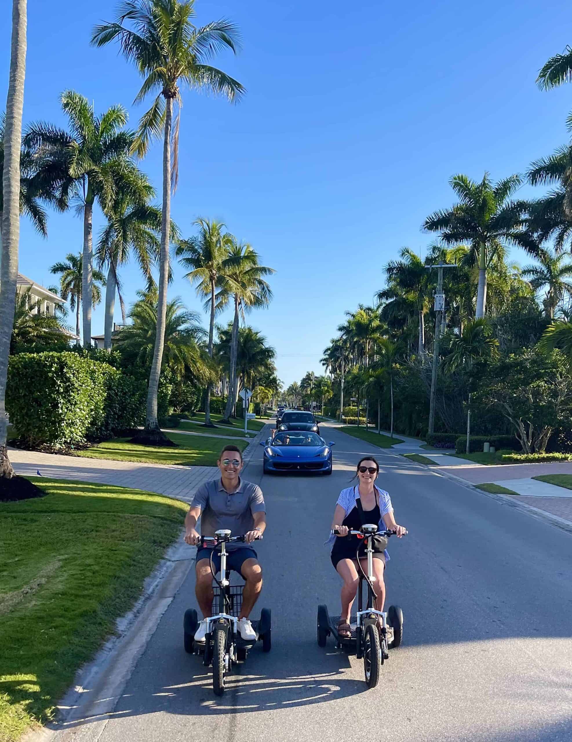 Side view of a smiling couple riding electric tricycles on a sunny street lined with palm trees in Naples, Florida, promoting pedal bike rentals for outdoor leisure and exploration.