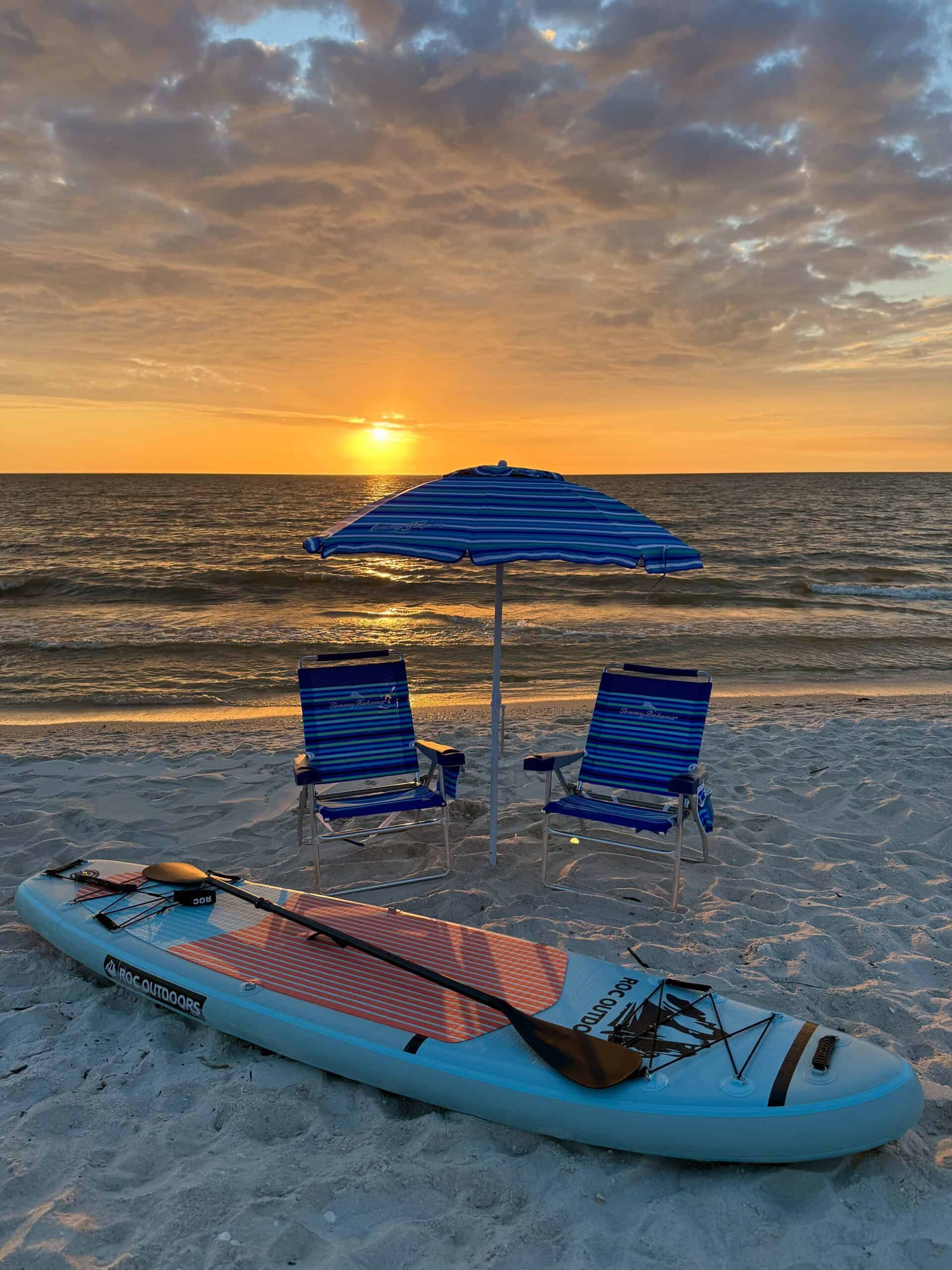 Relaxing beach scene with a paddleboard, two lounge chairs, and a beach umbrella during sunset, perfect for beach chairs and water sports rentals.