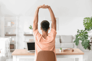 Relaxed woman stretching at home, sitting at a desk with a laptop, natural light, and green plants in background.