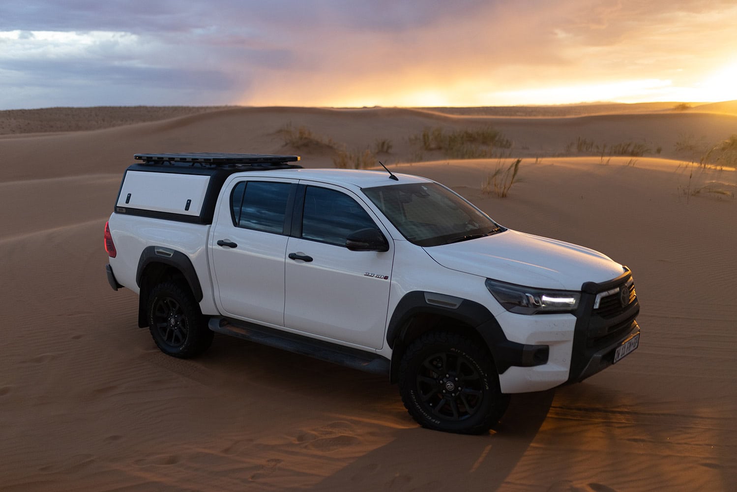Stainless steel canopy on a white pickup truck driving through desert sands at sunset.