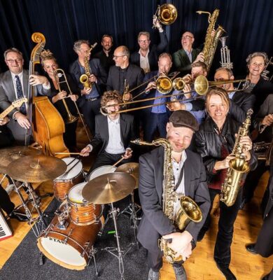 Een enthousiast orkest van jazz- en soulmusici met verschillende instrumenten, including gitaar, drums, saxofon, trompet en keyboard, op een podium met donkerblauw gordijn.