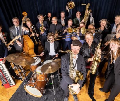 Een enthousiast orkest van jazz- en soulmusici met verschillende instrumenten, including gitaar, drums, saxofon, trompet en keyboard, op een podium met donkerblauw gordijn.