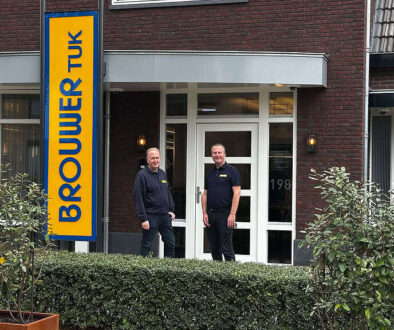 Bedrijfsgebouw van RTV SLOS met twee medewerkers voor de ingang, signaalbord met de naam, moderne gevel, personeel in uniforme kleding, lokale omroep, televisiestudio, radiostation, professionele uitstraling, communicatie, media-support, nieuwsvoorziening.