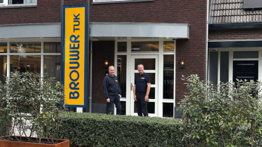 Brouwer Tuk - Foto Dirk Brans Bedrijfsgebouw van RTV SLOS met twee medewerkers voor de ingang, signaalbord met de naam, moderne gevel, personeel in uniforme kleding, lokale omroep, televisiestudio, radiostation, professionele uitstraling, communicatie, media-support, nieuwsvoorziening.