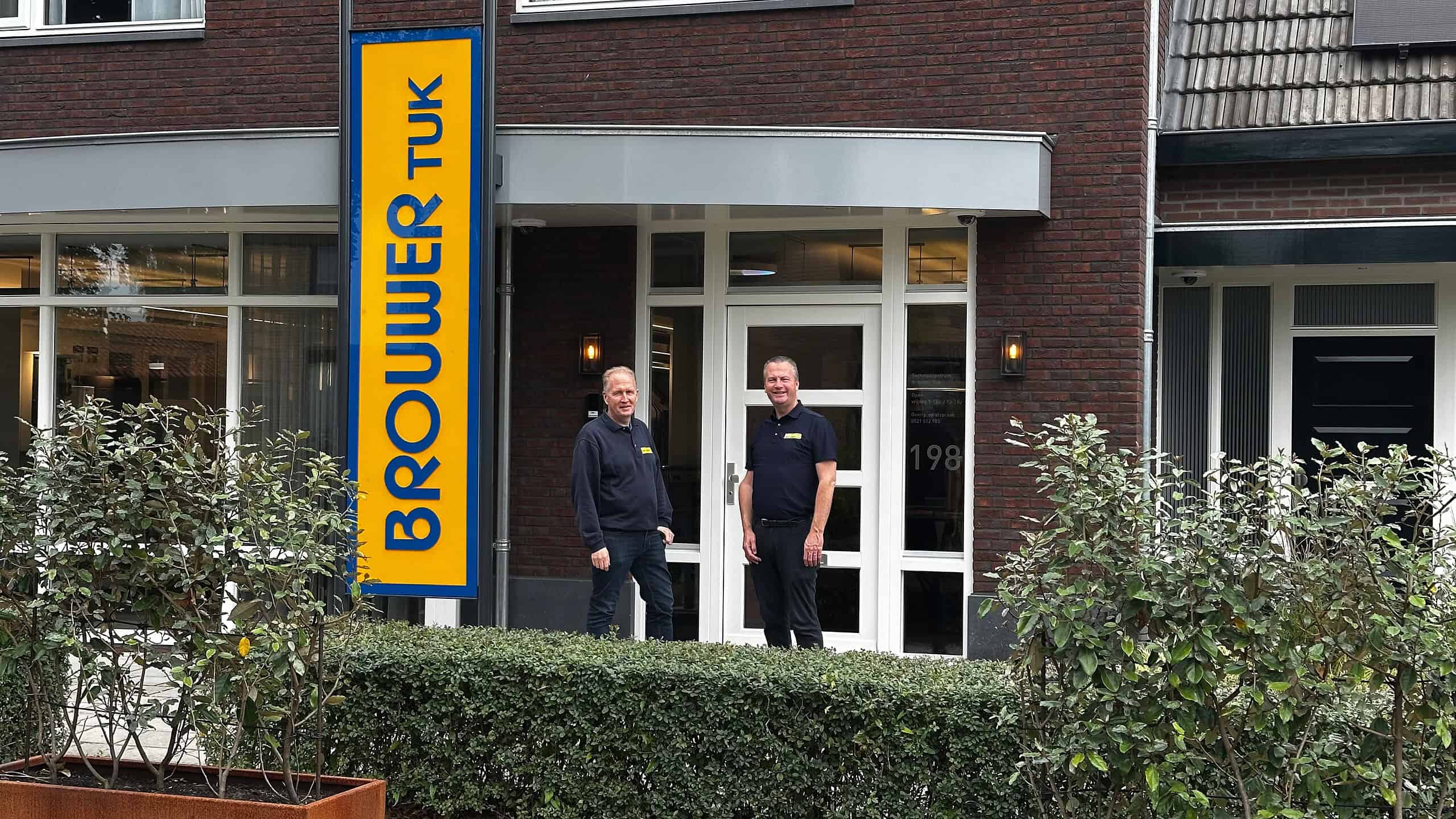 Bedrijfsgebouw van RTV SLOS met twee medewerkers voor de ingang, signaalbord met de naam, moderne gevel, personeel in uniforme kleding, lokale omroep, televisiestudio, radiostation, professionele uitstraling, communicatie, media-support, nieuwsvoorziening.