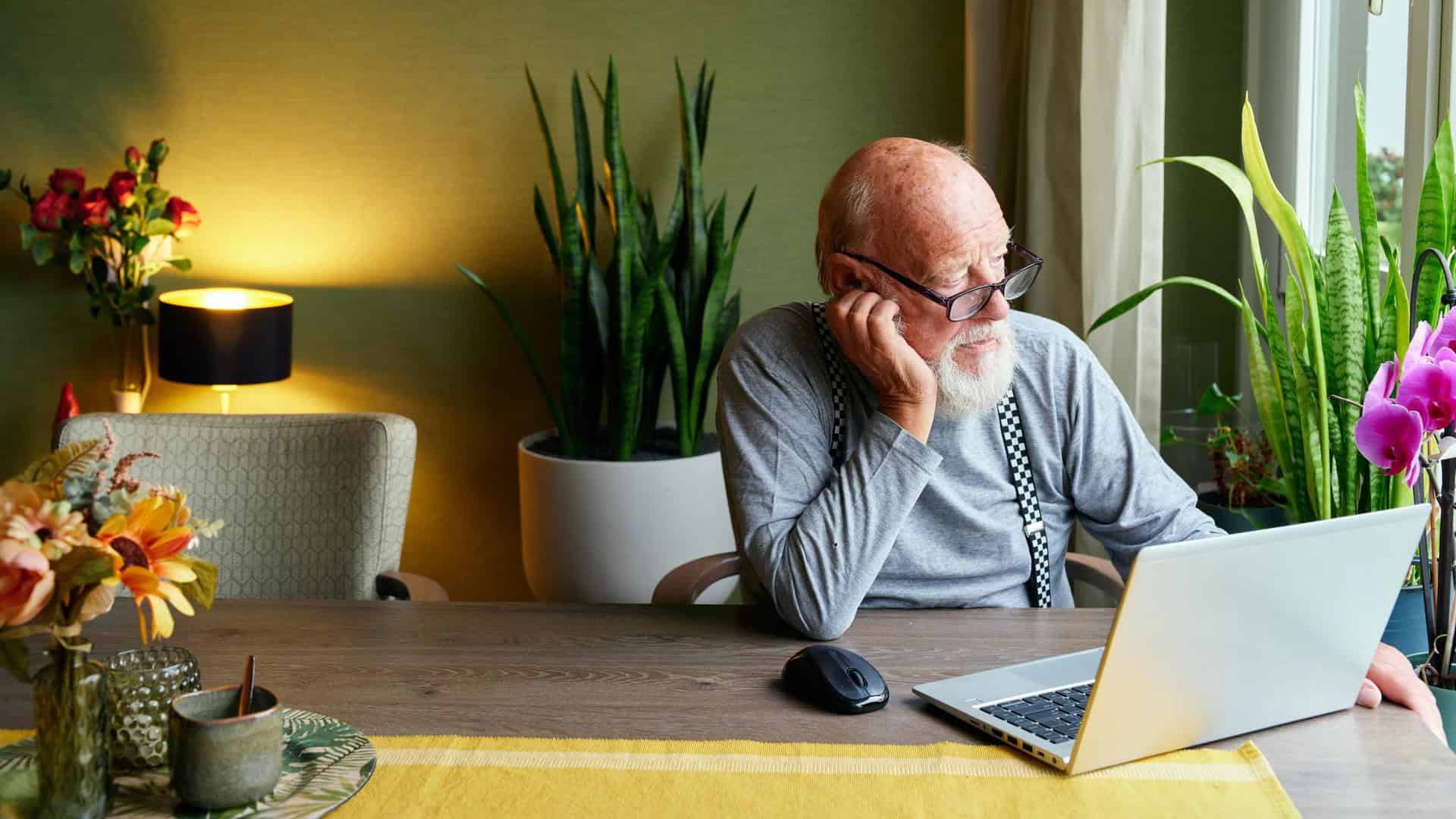 Knusse woonkamer met oudere man workend op laptop, omringd door groene planten, bloemen en sfeervol licht, perfect voor een gezellige en rustgevende sfeer.