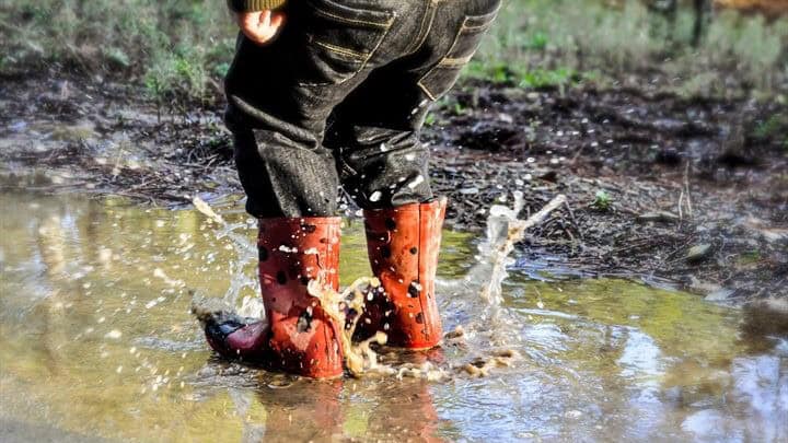 Kind met oranje rubberlaarzen waden door modderig water in een natuurgebied.