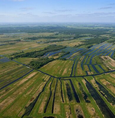 Luchtfoto van de uitgestrekte waterrijke landbouwgebieden in de omgeving van RTV SLOS, met diverse weilanden, sloten en irrigatiekanalen in een weids landschap.