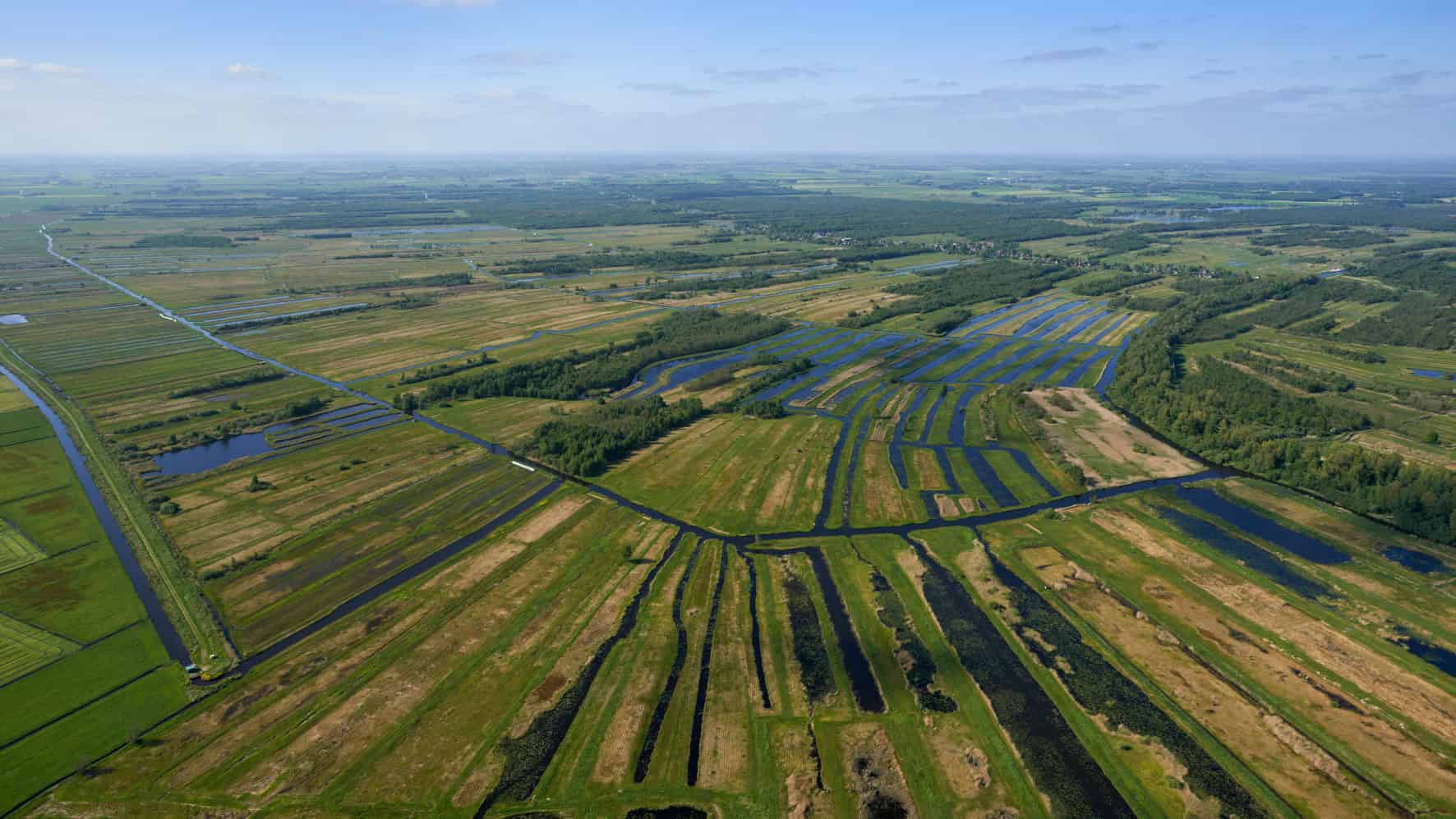 Luchtfoto van de uitgestrekte waterrijke landbouwgebieden in de omgeving van RTV SLOS, met diverse weilanden, sloten en irrigatiekanalen in een weids landschap.