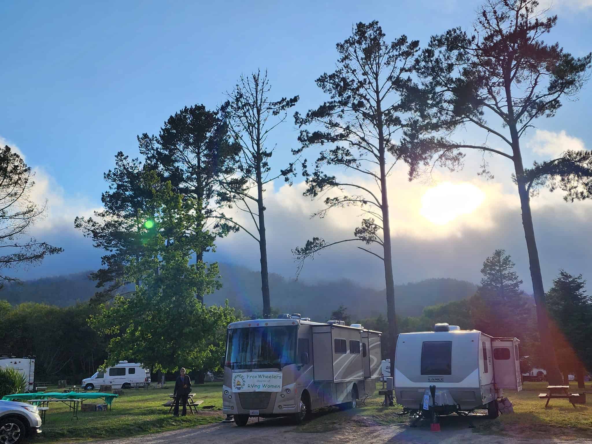 RVers parked in a scenic outdoor setting with tall trees and mountains in the background, enjoying a peaceful camping experience with sunlight filtering through the sky.