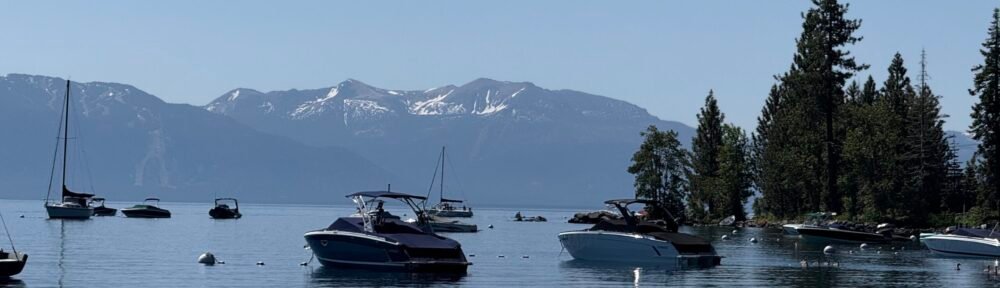 Boats floating on calm lake with mountain backdrop and pine trees, peaceful summer scenery for outdoor recreation and water sports.
