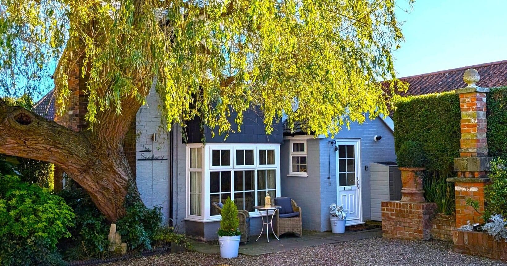 Cottage-style home with a bay window, wicker seating, potted plants, and “The Saddlery” on the wall beneath a large tree.