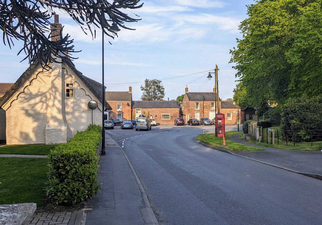 The Square in North Thoresby. A thatched house on the left and old buildings ahead. The memorial gardens to the right and a traditional red telephone box.