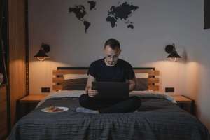 a man using a laptop while sitting on a bed.Digital Nomad Meaning